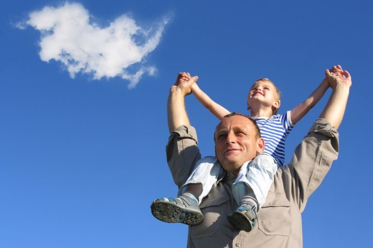 Grandfather holding grandson on his shoulders with arms up and smiling on a bright blue-sky day